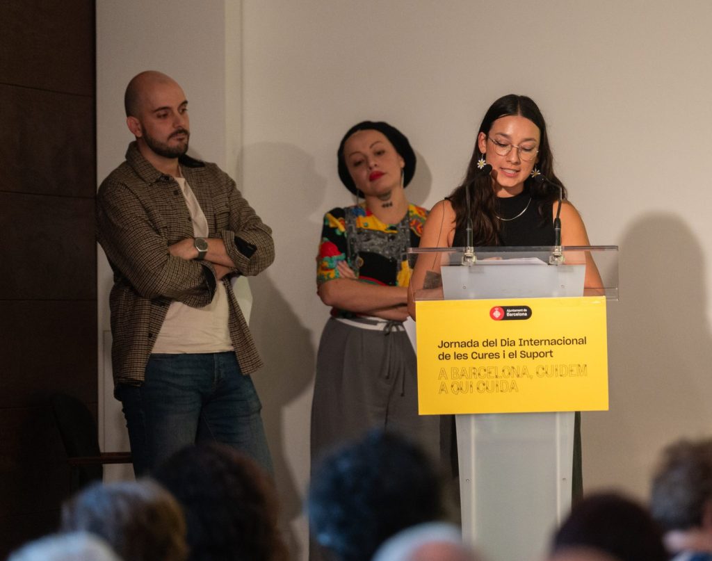 Joves cuidadores. Un dels membres del grup de treball parla des d'un faristol amb dos micròfons, mentre dues dones l'escolten des del darrere. L'home porta una camisa de quadres oberta i una samarreta clara a sota. Una de les dones porta ulleres, una samarreta negra sense mànigues i sosté uns papers. L'altra dona porta una samarreta amb estampat colorit, un mocador negre al cap i també sosté papers. Al faristol hi ha un cartell groc amb el logotip de l'Ajuntament de Barcelona i el text: 'Jornada del Dia Internacional de les Cures i el Suport. A Barcelona, cuidem a qui cuida